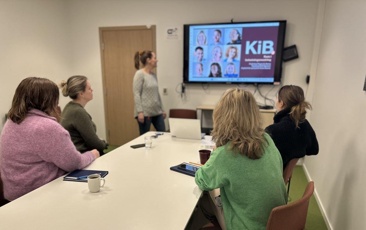 Fire personar sit rundt eit bord. Ein person viser noko på ein skjerm på veggen - Klikk for stort bilete